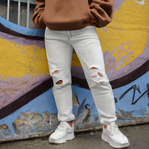 Người mặc giày chunky màu trắng, quần jean flare high-waist màu sáng và áo hoodie màu nâu đứng trước tường graffiti.
